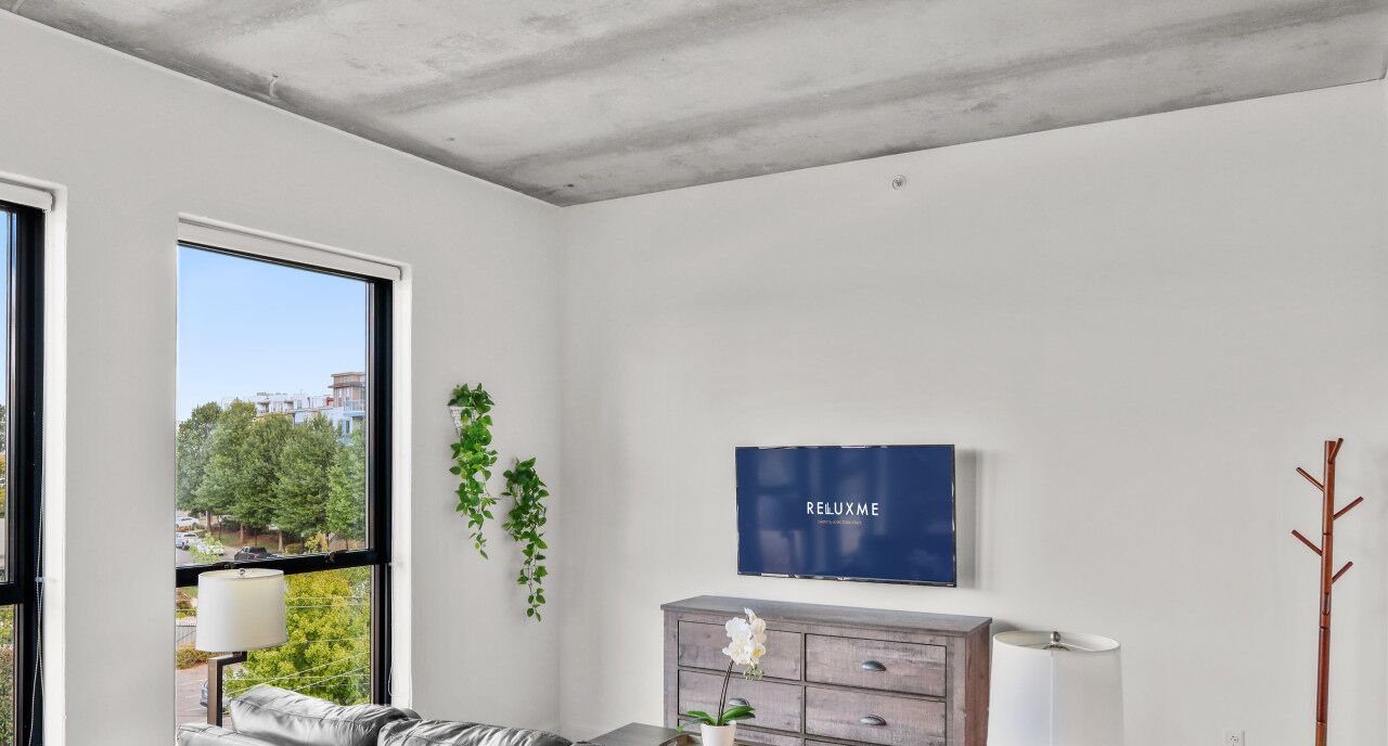 A modern living room with a large window, a flat-screen TV, a grey dresser, and a plant.