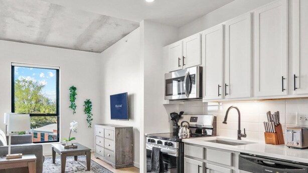 A modern kitchen with white cabinets, a microwave, stove, and a dishwasher. There is a window with a view of trees and a blue sign on the wall. - Reluxme|European Flat Near Uptown King Bed + Views (Charlotte)
