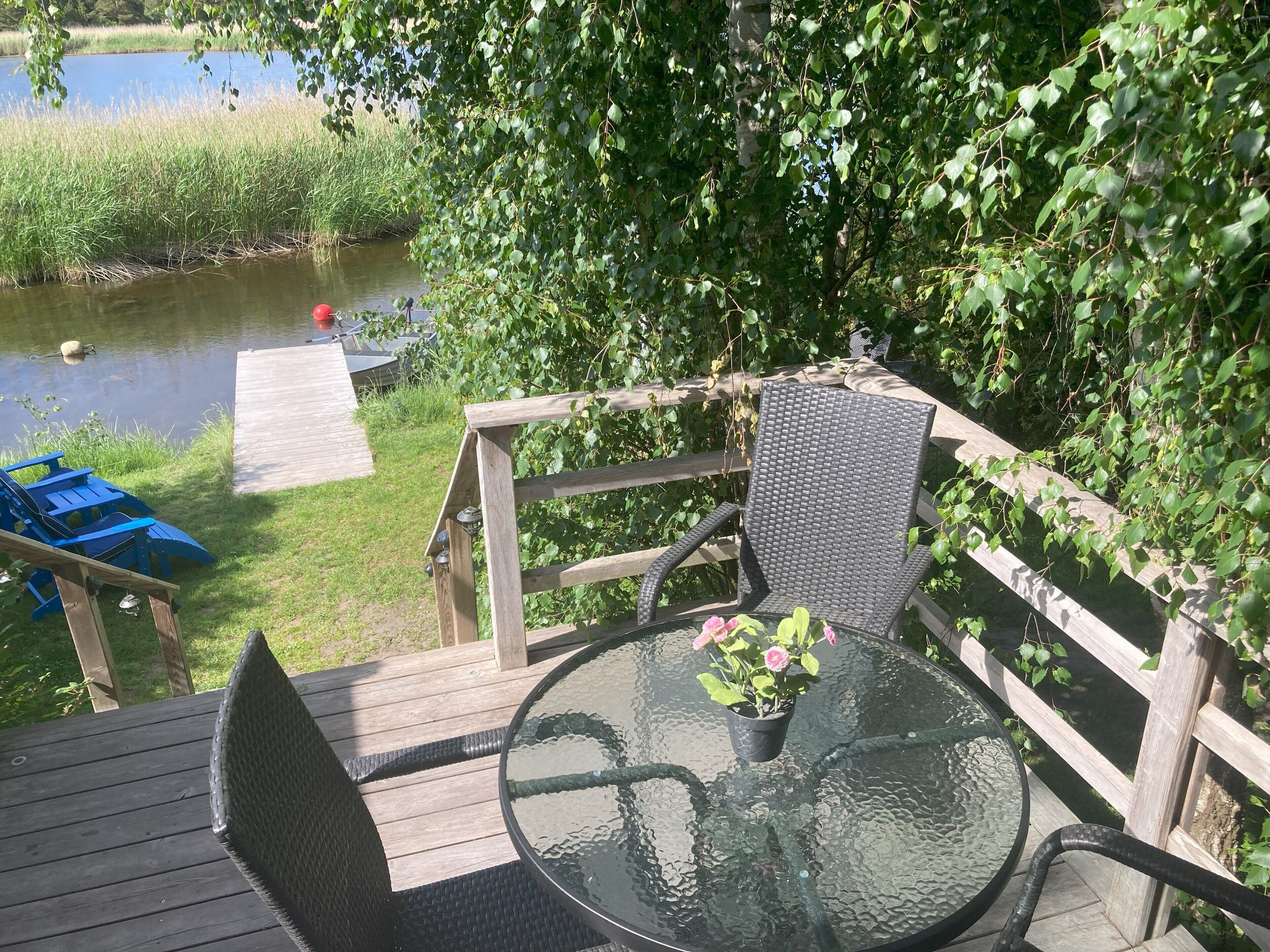 Una terraza de madera con una mesa y sillas de vidrio, con vista a una masa de agua rodeada de vegetación.
