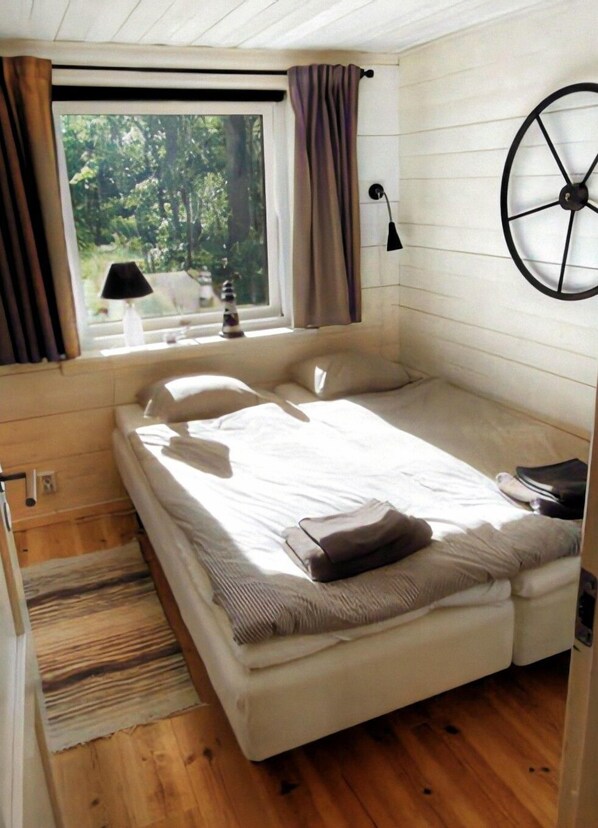 A small, cozy bedroom with a single bed, a window with curtains, a lamp, and a wall clock. - Sjöstugan (Rockneby)
