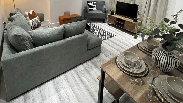A modern living room with a grey sofa, a dining table set with plates and glasses, a flat-screen TV on a wooden stand, and a floor lamp.
