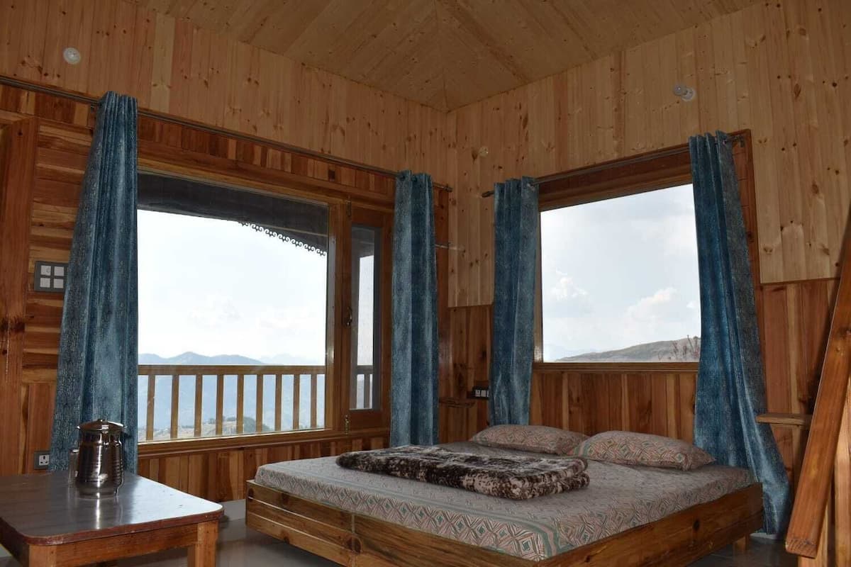 A wooden cabin bedroom with a bed, a table, and large windows with curtains.