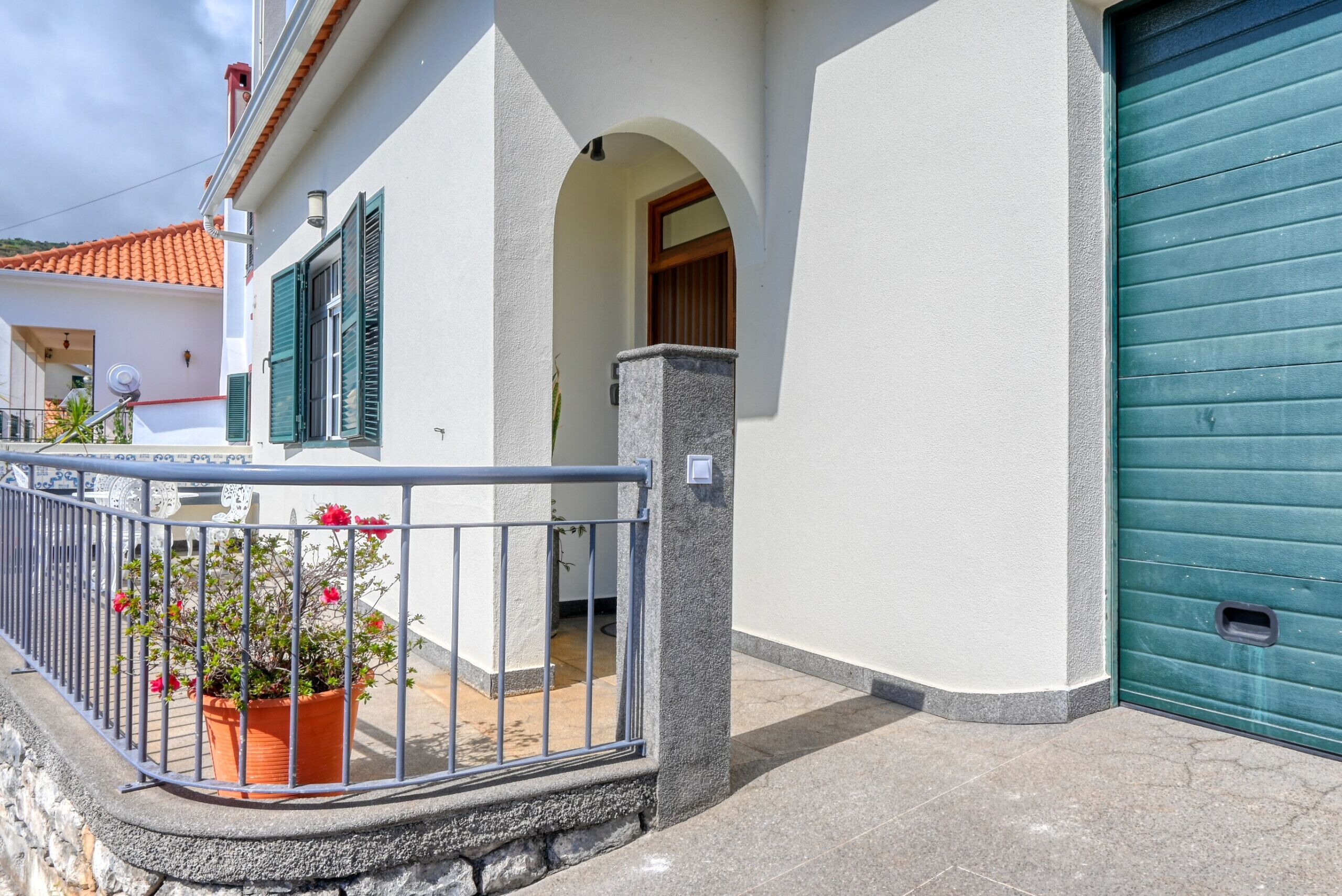 A building with a green shuttered garage, a potted plant with red flowers, and a stone pillar.