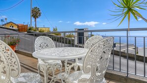 En balkon med detaljerede hvide metalstole og et bord, med udsigt over et kystlandskab med et palmetræ og en bygning i baggrunden.