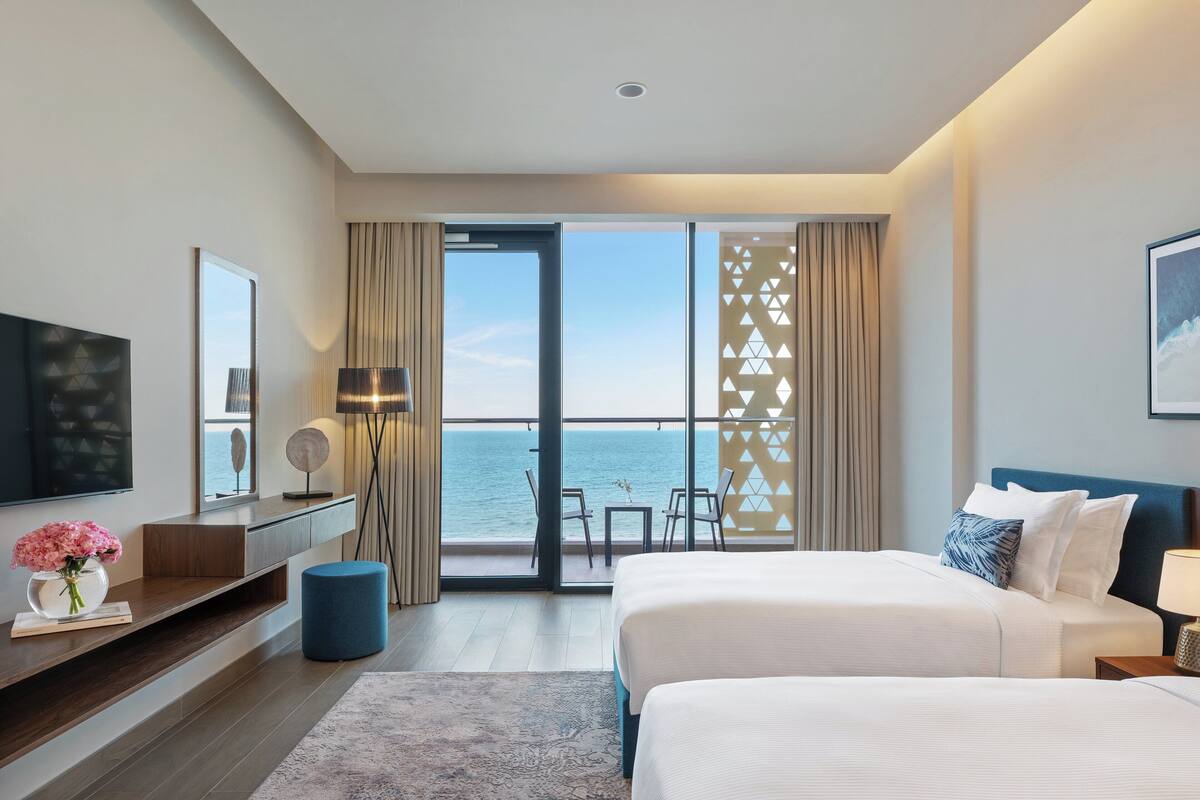 A modern hotel room with a large bed, a TV, a desk with a lamp, and a view of the sea through a glass door.