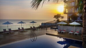 Piscina infinita en la azotea con sillones de descanso y sombrillas, con vista al océano al atardecer.
