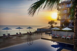 Piscina infinita no terraço com cadeiras de descanso e guarda-sóis, com vista para o oceano ao pôr do sol.