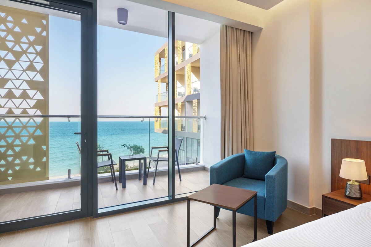 Habitación de hotel con balcón con vistas al mar, un sofá, una mesita y una mesita de noche con lámpara.