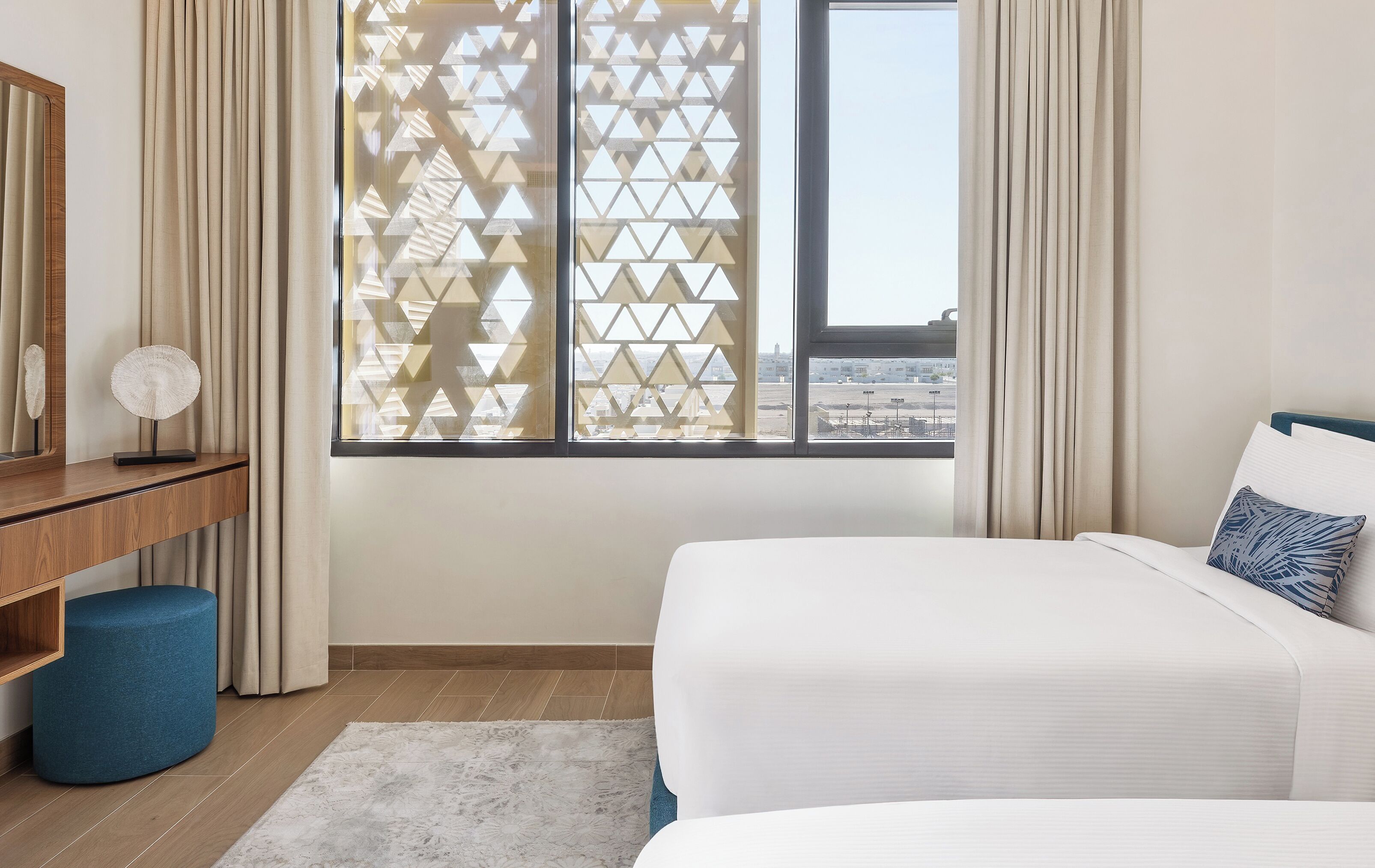 Une chambre d’hôtel comprenant un lit, un bureau, une chaise, un petit pouf rond, un tapis blanc et une fenêtre donnant sur la ville.