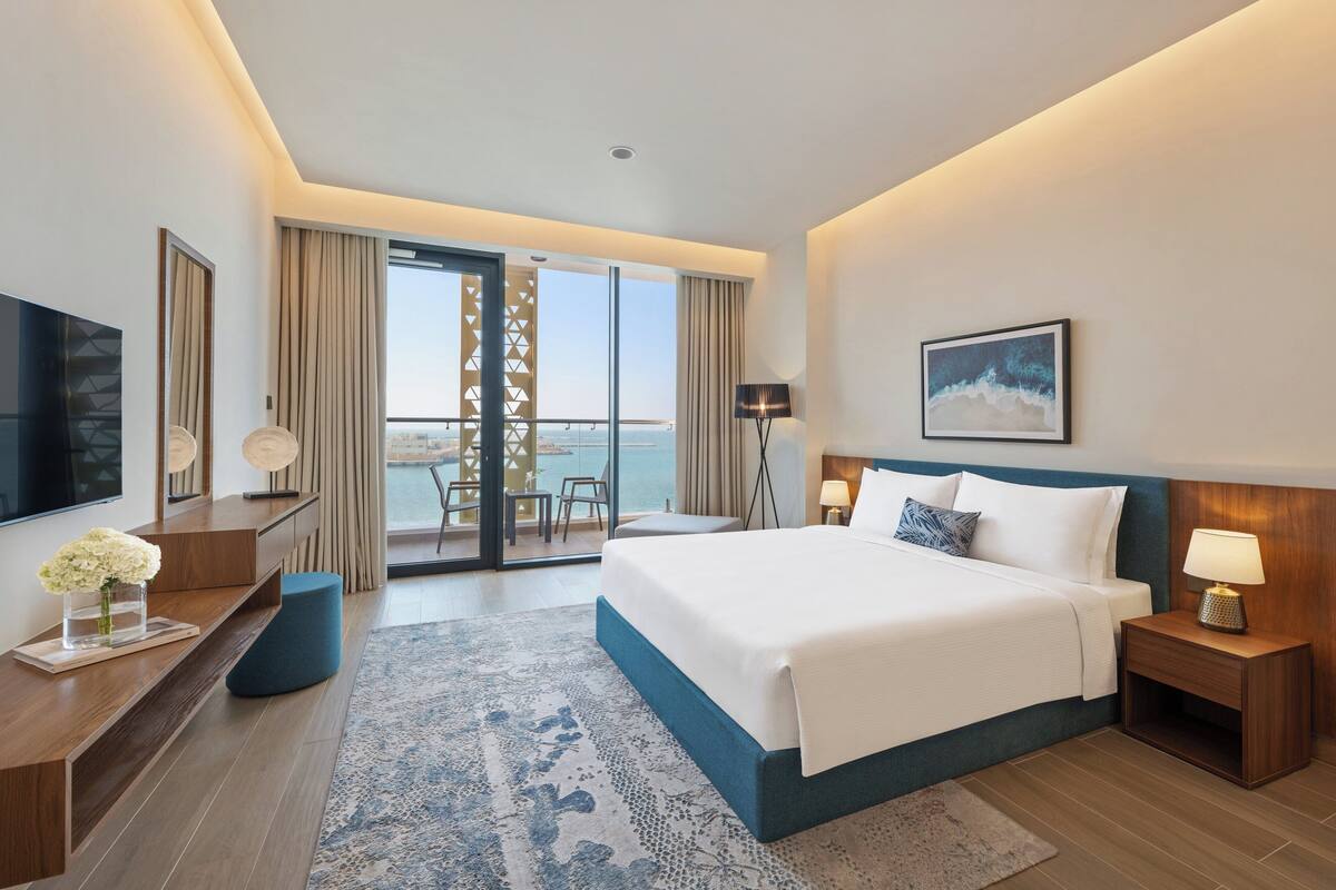 A modern hotel room with a large bed, a TV, a desk, and a balcony with a view of the sea.