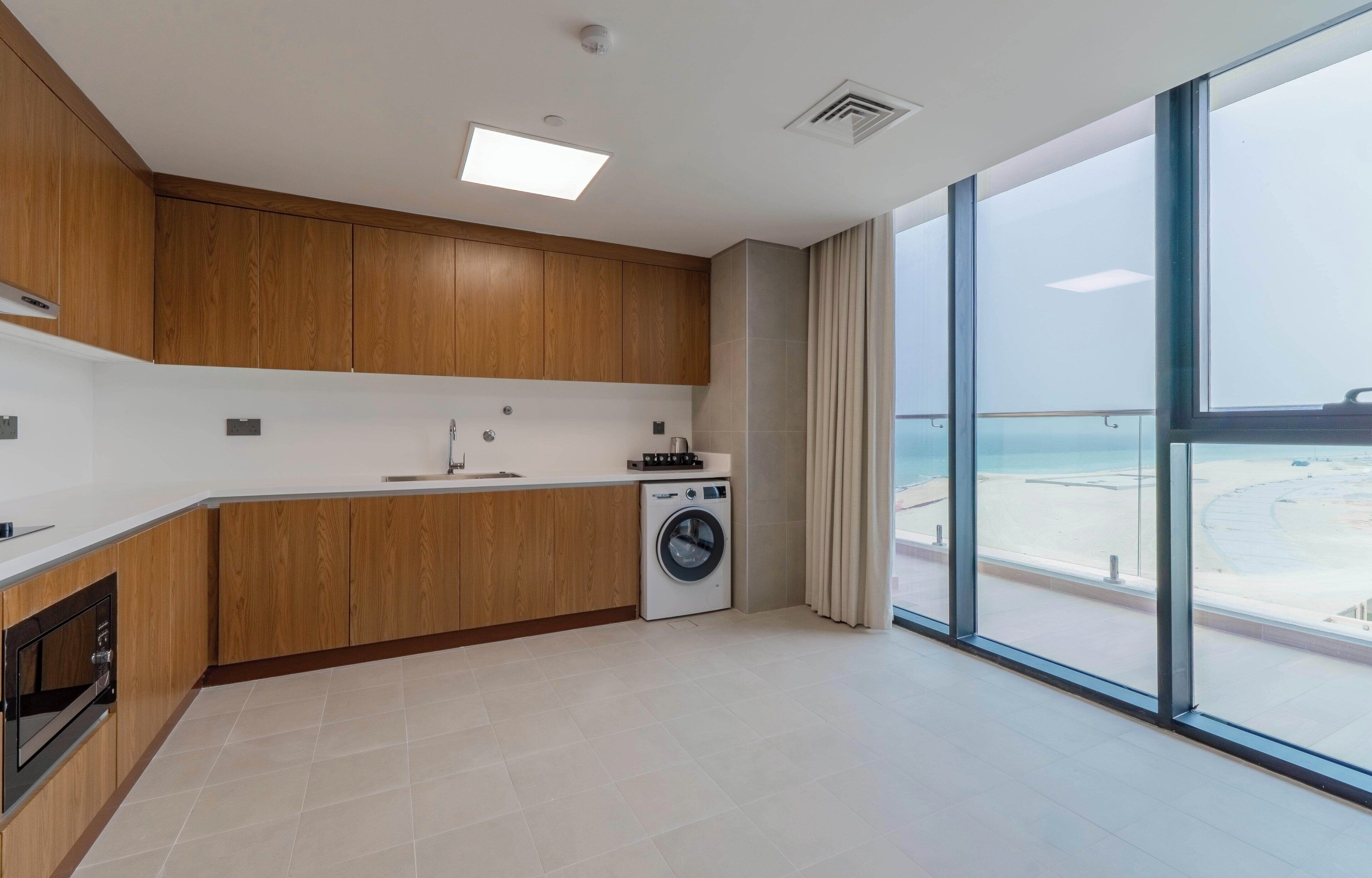 Una cocina moderna con armarios de madera, lavadora integrada y un amplio ventanal con vistas a la playa.