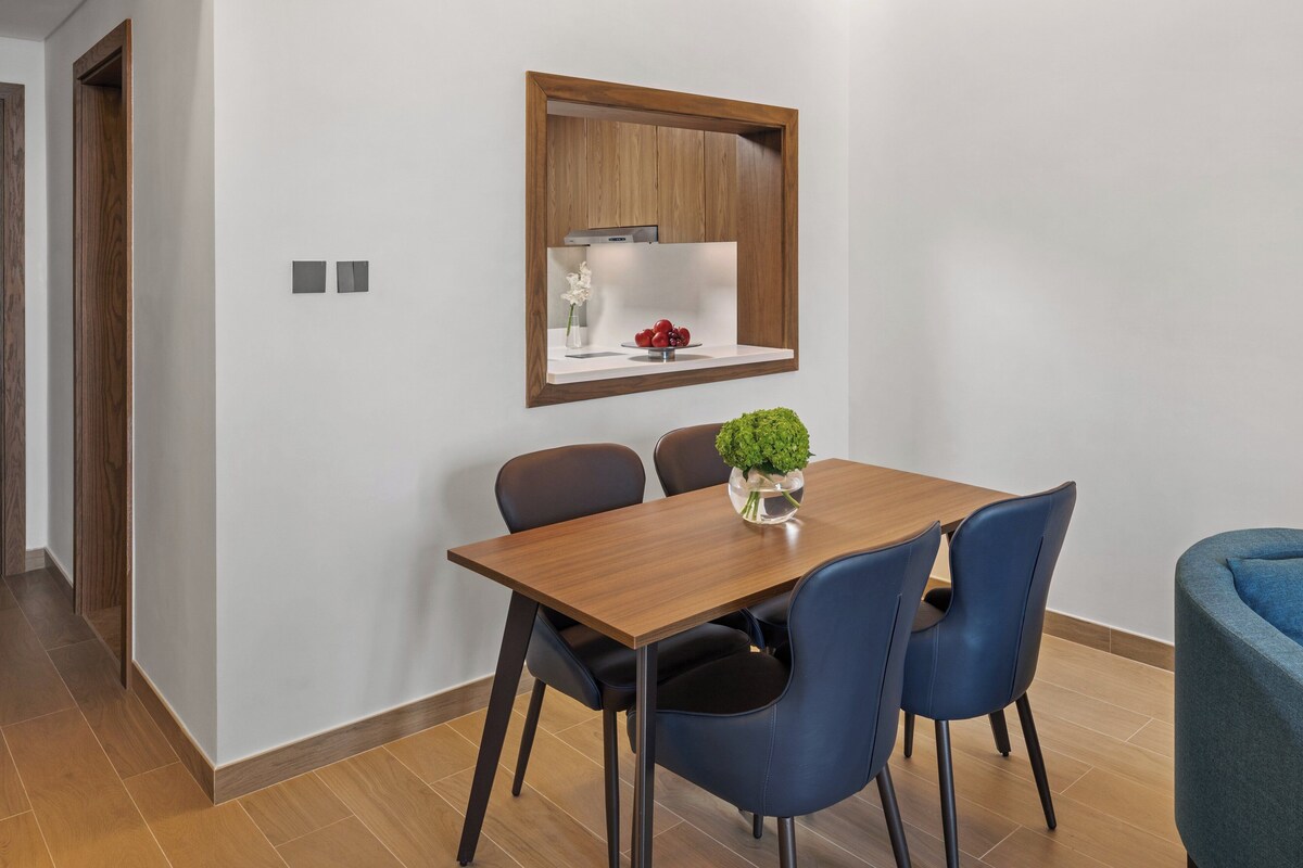 Un comedor con una mesa de madera, cuatro sillas, una planta pequeña en maceta y un estante en la pared con un jarrón y fruta.
