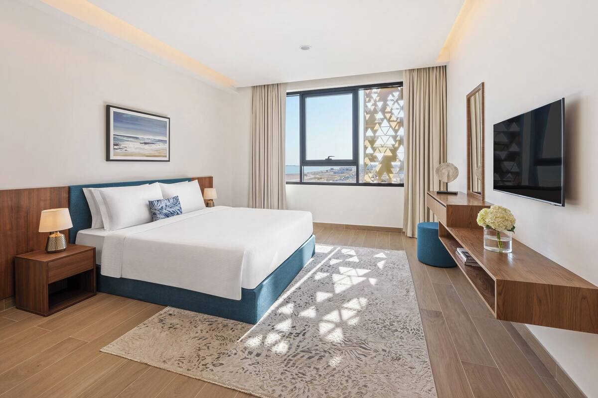A modern hotel room with a large bed, a TV, a desk, and a view of the beach.