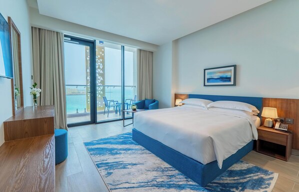 A hotel room with a large bed, a desk, a chair, and a balcony with a view of the sea.