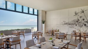 Un comedor con mesas dispuestas para recibir a los invitados, ventanas amplias que ofrecen vistas al mar y un mural que representa una escena costera.