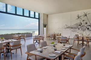 Uma sala de jantar com mesas preparadas para hóspedes, grandes janelas com vista para o mar e um mural retratando uma paisagem costeira.