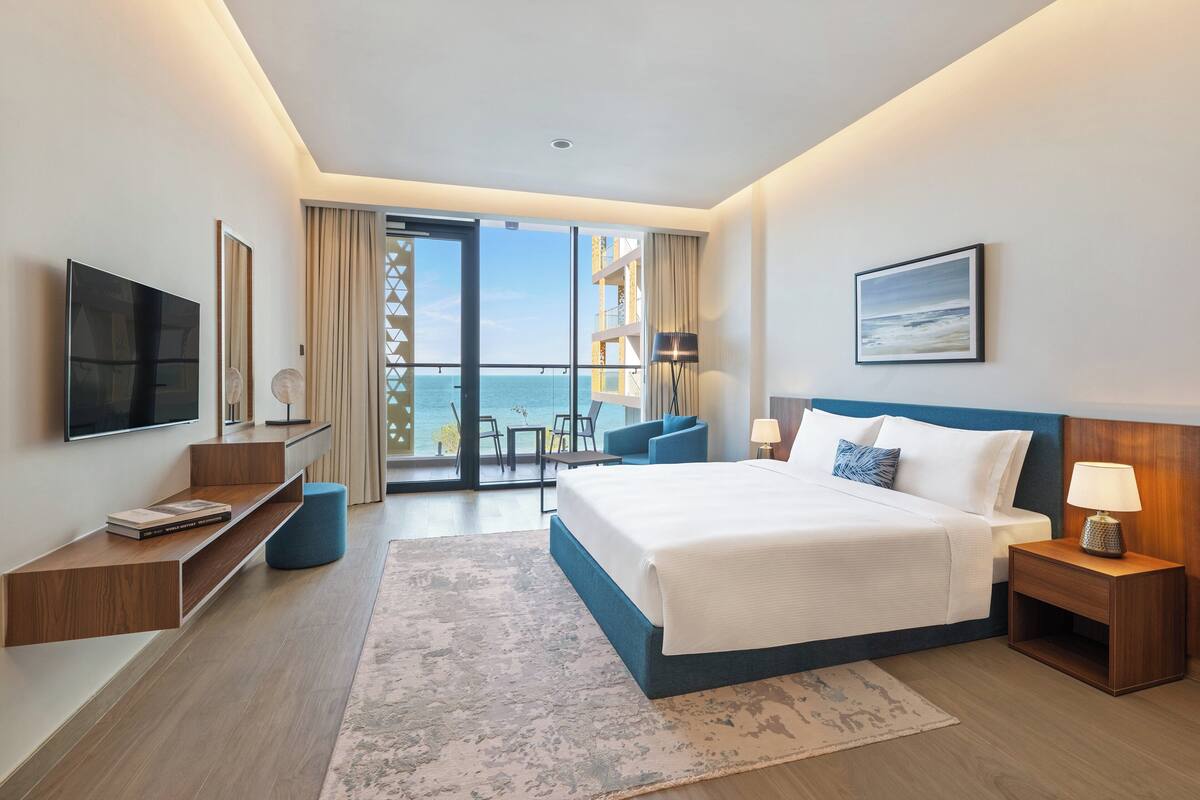 A modern hotel room with a large bed, a TV, a desk, and a balcony with a view of the sea.