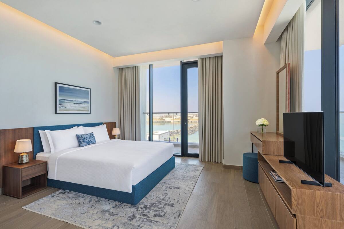 A modern hotel room with a large bed, a flat-screen TV, a wooden dresser, and a view of the city through large windows.