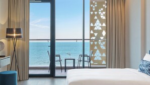 Habitación de hotel con una ventana grande que ofrece vista al mar, una cama, un escritorio con lámpara y una pequeña mesa con sillas.