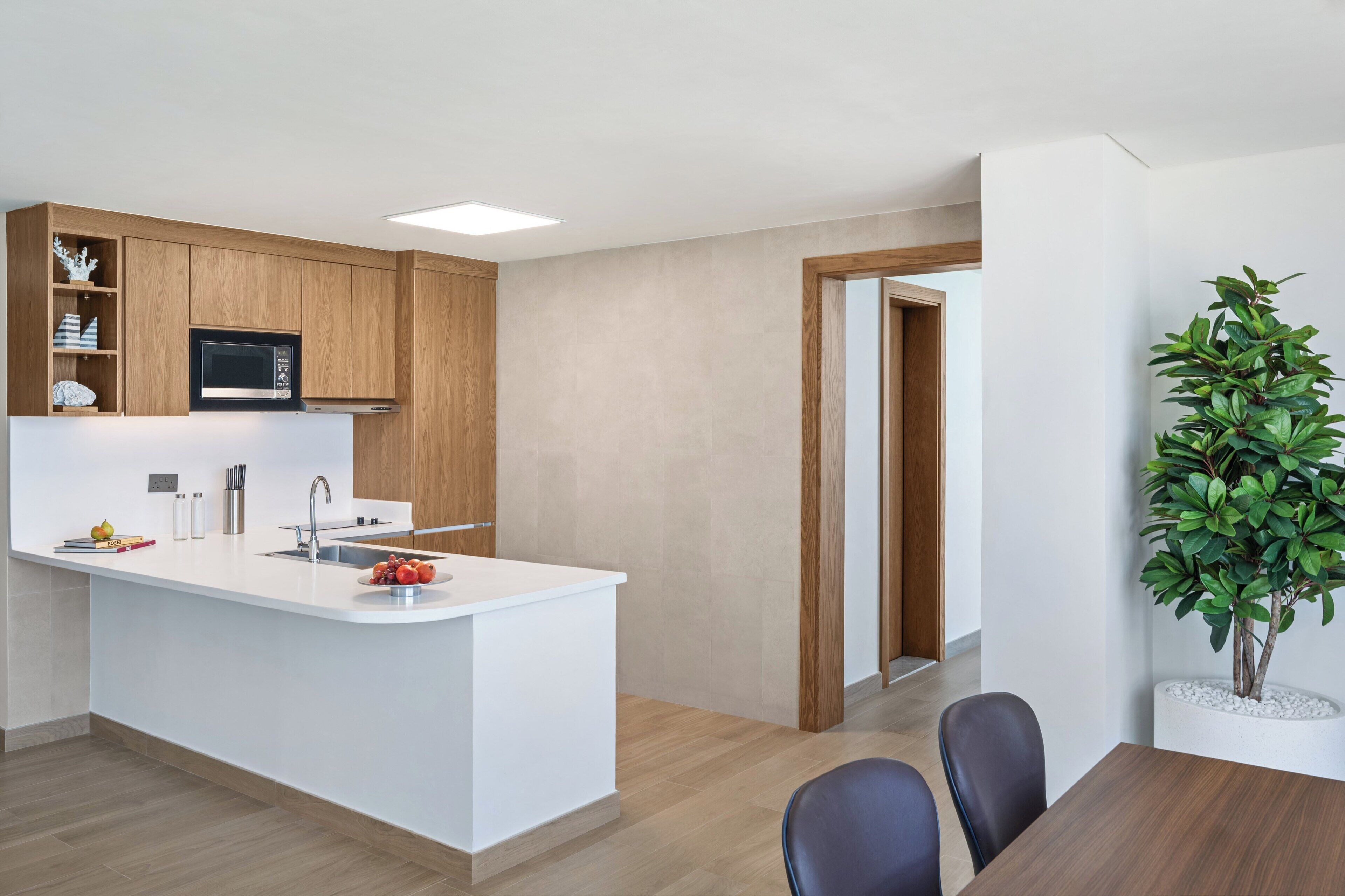 A modern kitchen with wooden cabinets, a white countertop, and a built-in microwave. There is a dining area with a table and chairs, and a potted plant on the right.
