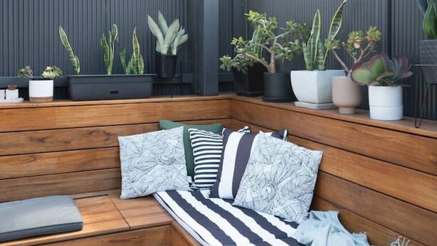 Un banc en bois agrémenté de coussins et recouvert d’une couverture, entouré de plantes en pot.