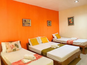 Une chambre d’hôtel avec trois lits simples, chacun doté d’un matelas blanc et d’un oreiller. Deux tableaux encadrés ornent les murs orange.