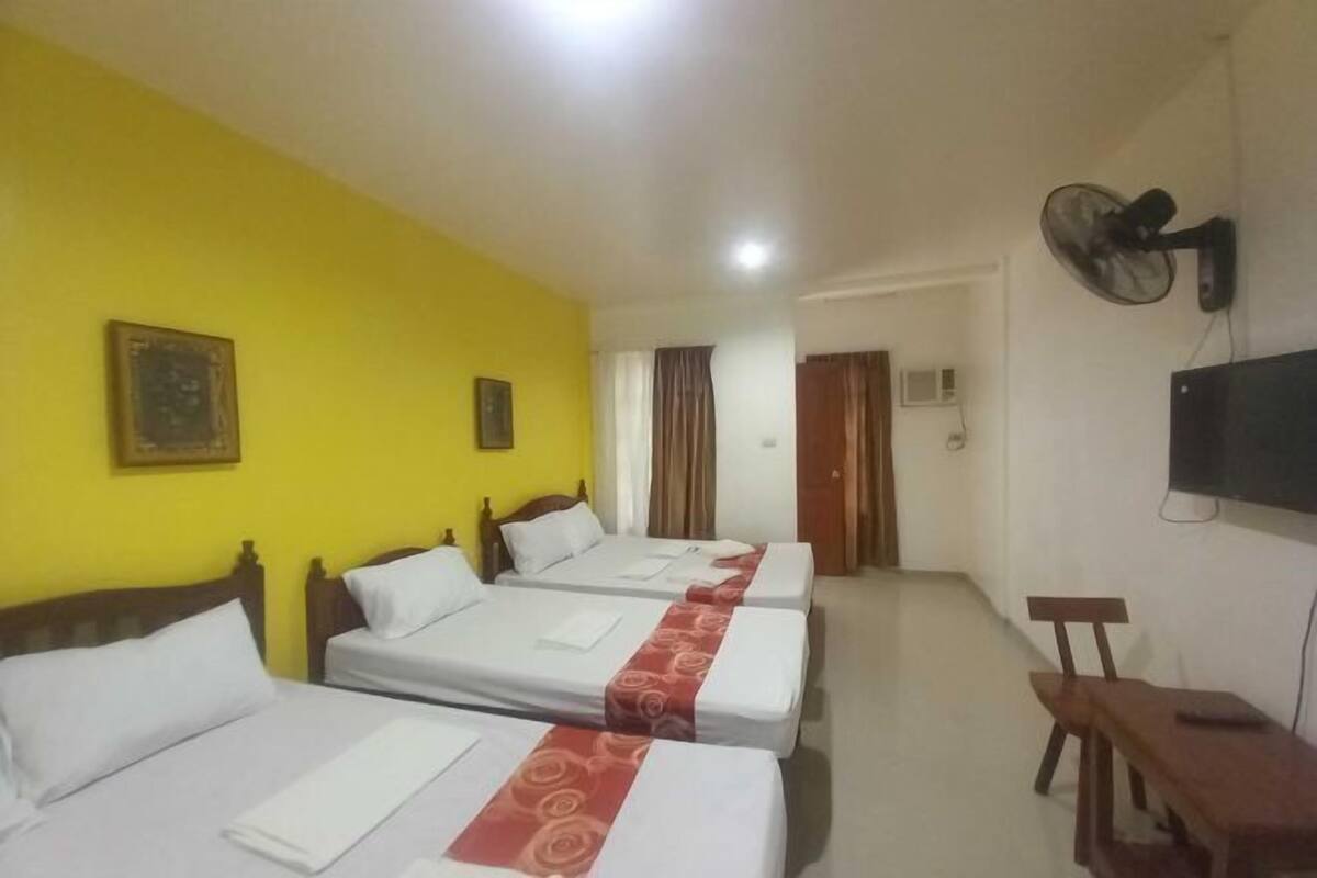 A hotel room with three beds, a TV, a fan, and a desk.