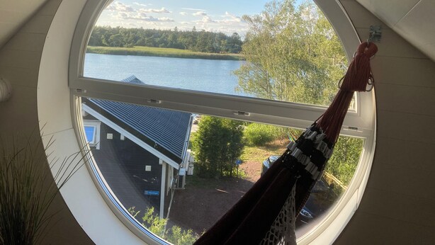 A round window with a view of a lake, trees, and a building with a dark roof. - Brygghuset (Rockneby)