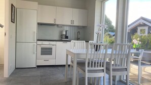 A modern kitchen with white cabinets, a built-in oven, and a coffee maker. There is a dining area with white chairs and a table, and a view of the outside through large windows. - Brygghuset (Rockneby)