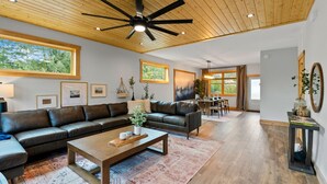 Un salón amplio con un gran ventilador de techo, piso de madera y vistas al exterior a través de amplios ventanales.