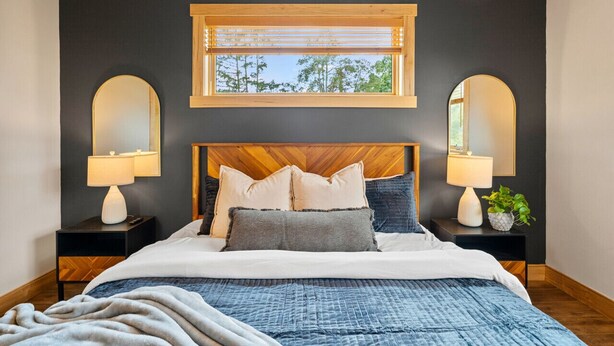 Un dormitorio con cabecera de madera, un ventanal con persianas, dos mesitas de noche con lámparas y una cama con muchos almohadones.