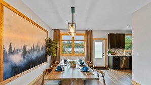 Un comedor con una mesa de madera dispuesta para seis personas, sillas a cada lado, un cuadro grande y vistas a través de una ventana.