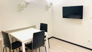 Een kamer met een witte tafel, vier zwarte stoelen, een flatscreen televisie aan de muur en twee wandlampen.