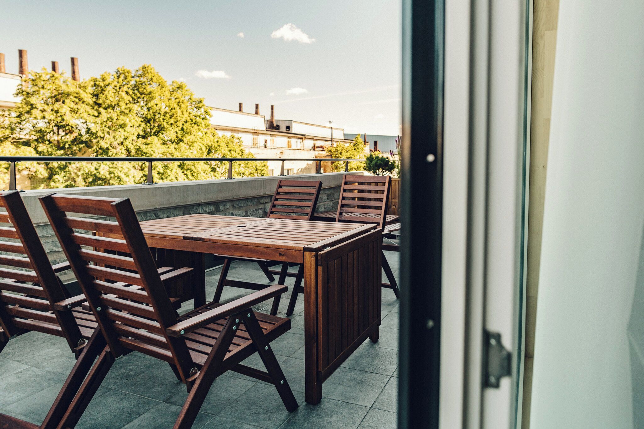 Eine Terrasse im Freien mit einem Holztisch und Stühlen, im Hintergrund Gebäude und Bäume.