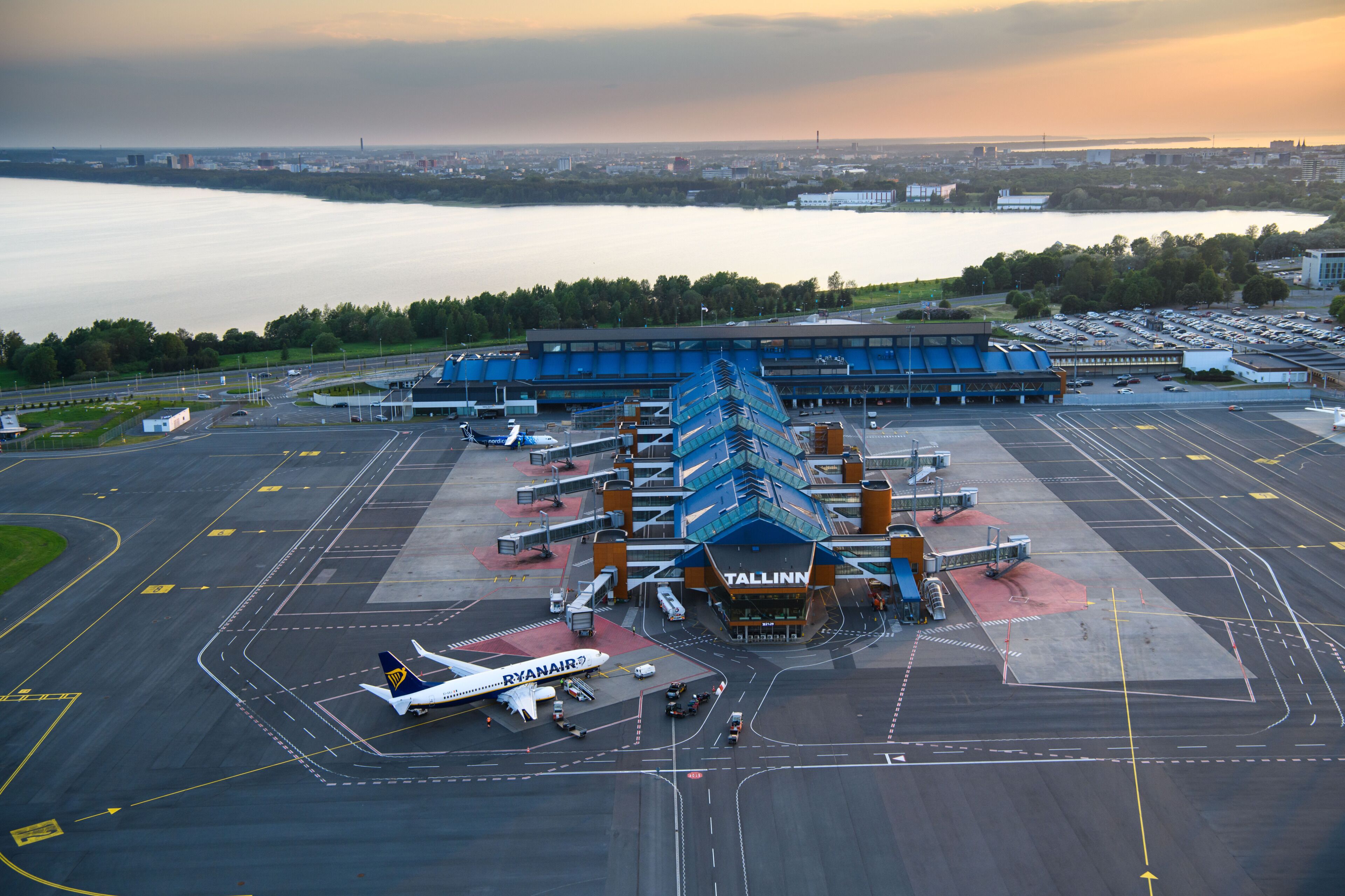 An airport terminal with a large building, parked airplanes, and a body of water in the background.