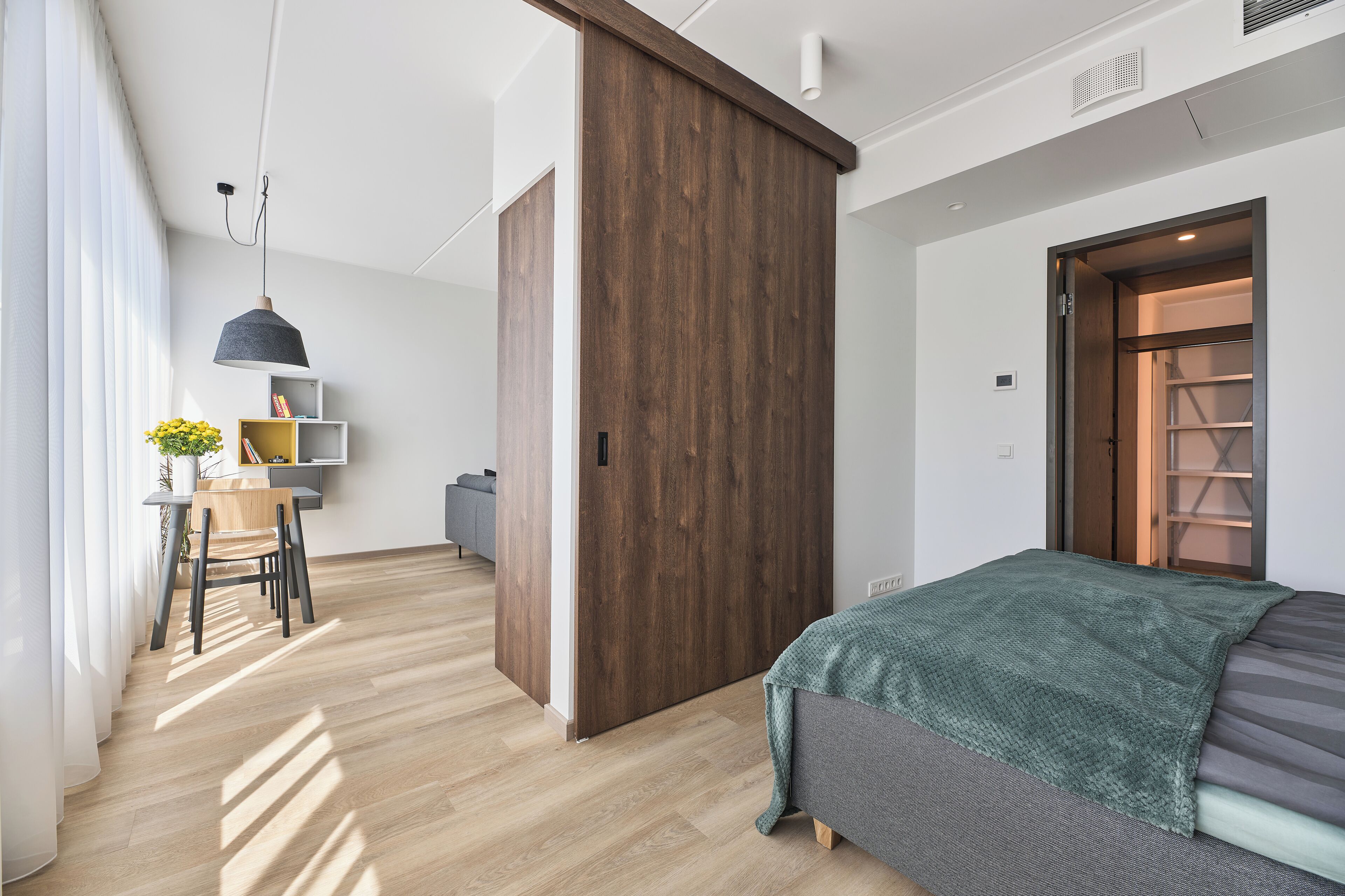 Quarto moderno com guarda-roupa de madeira, cama com manta verde e escrivaninha com cadeira e planta.
