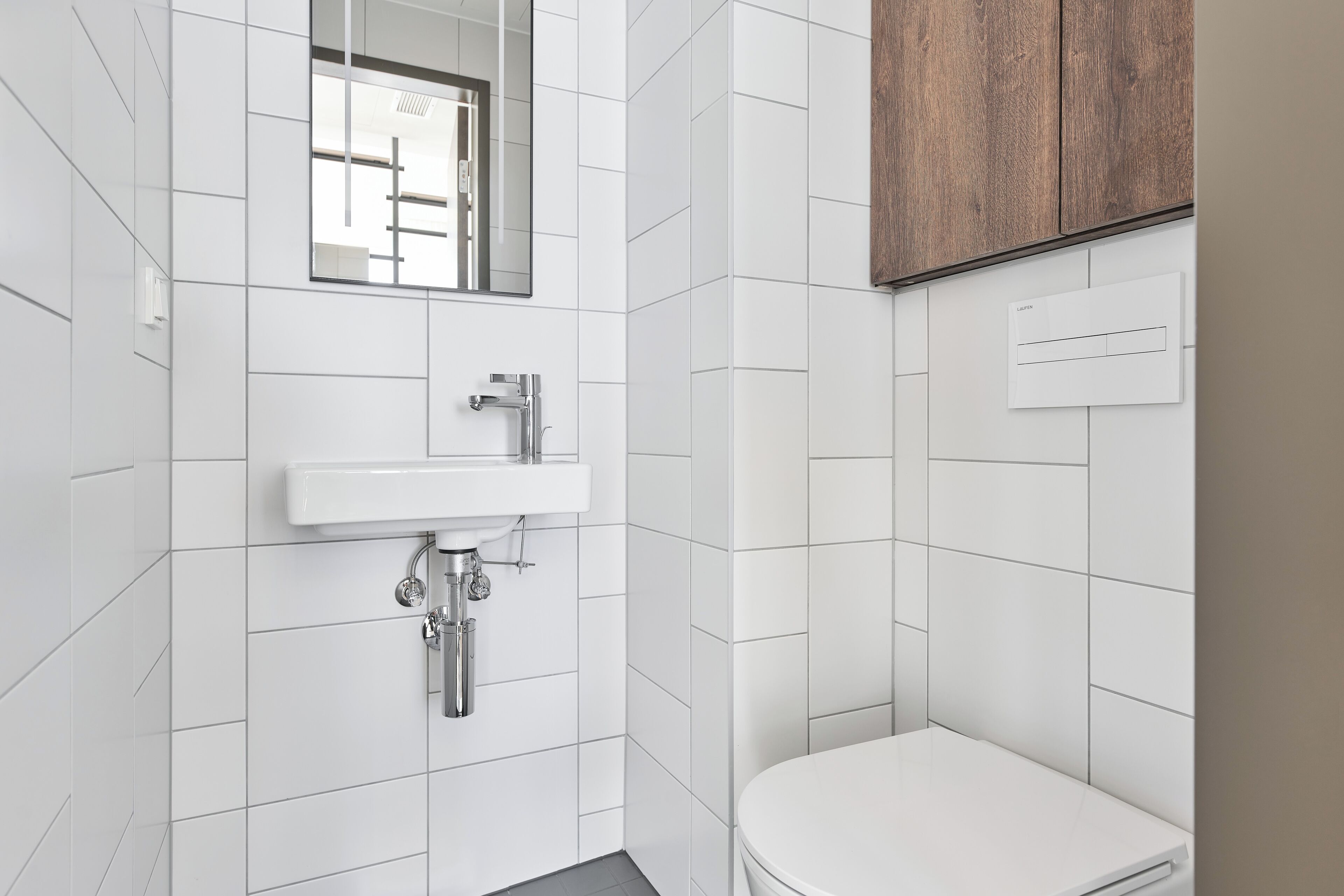 A modern bathroom with white tiles, a white sink, a mirror, and a wooden cabinet.