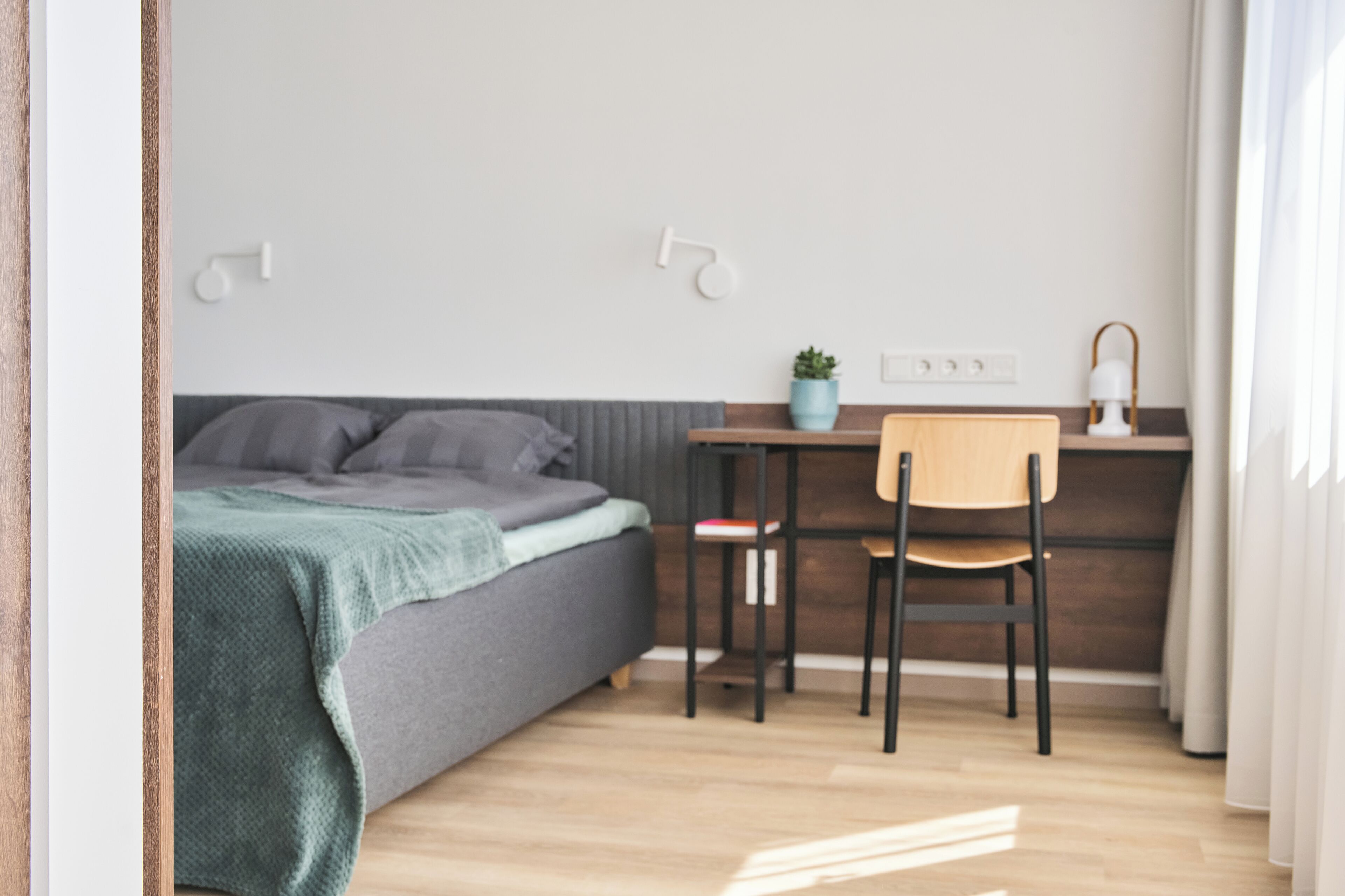 Ein Schlafzimmer mit Bett, Schreibtisch, Stuhl, einer Zimmerpflanze und einem Nachttisch.