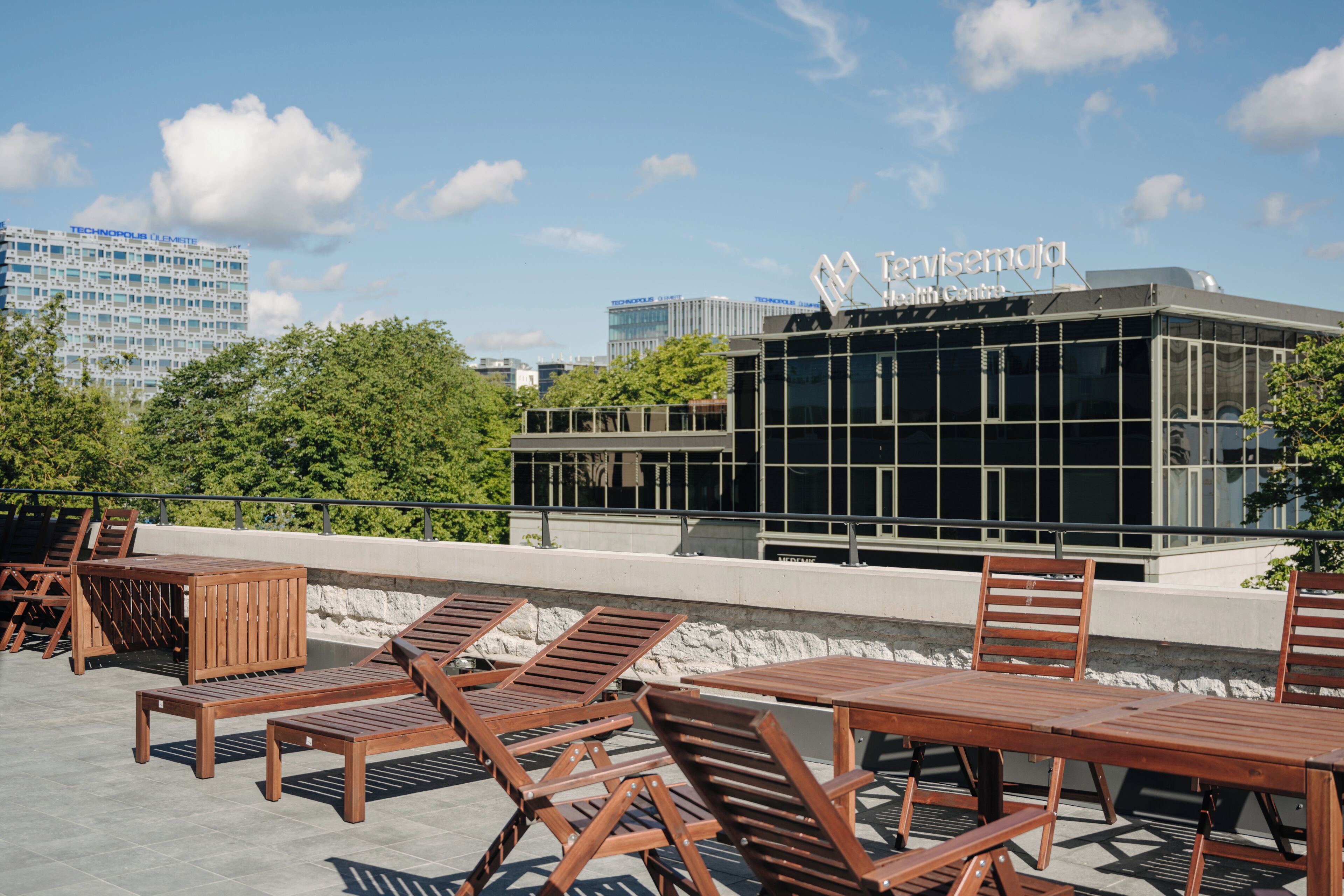 Una terraza en la azotea con sillones y mesas de madera, un edificio moderno con ventanas de vidrio y un cielo azul despejado.
