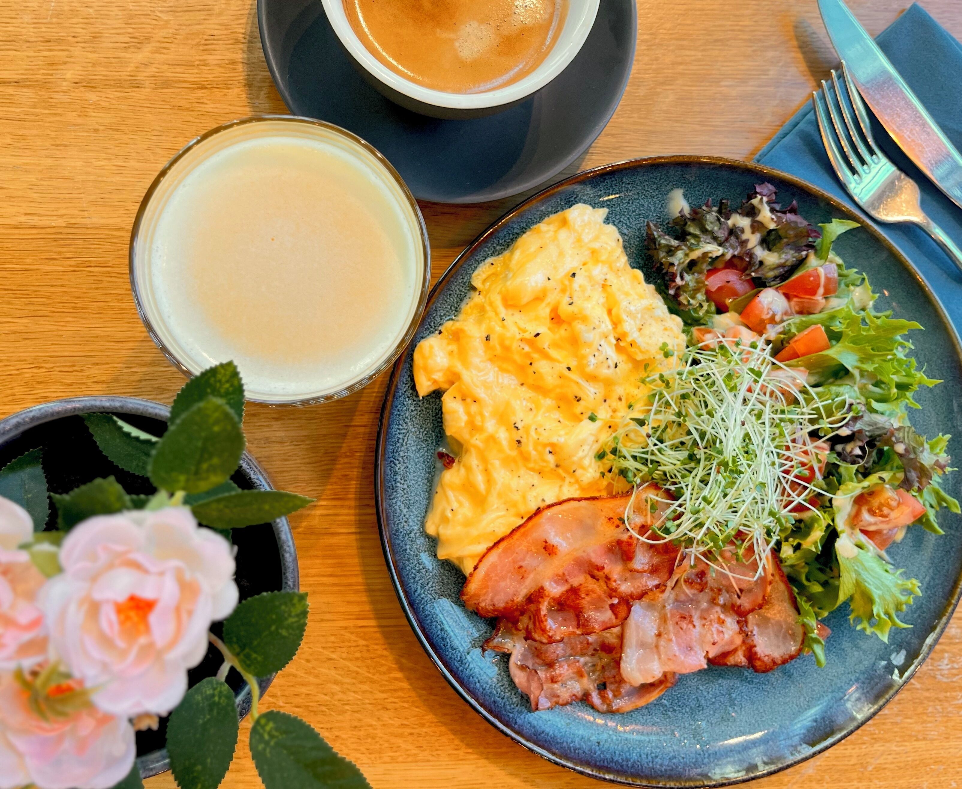 A plate of scrambled eggs, bacon, and a salad with sprouts, accompanied by a cup of coffee and a glass of milk on a wooden table.