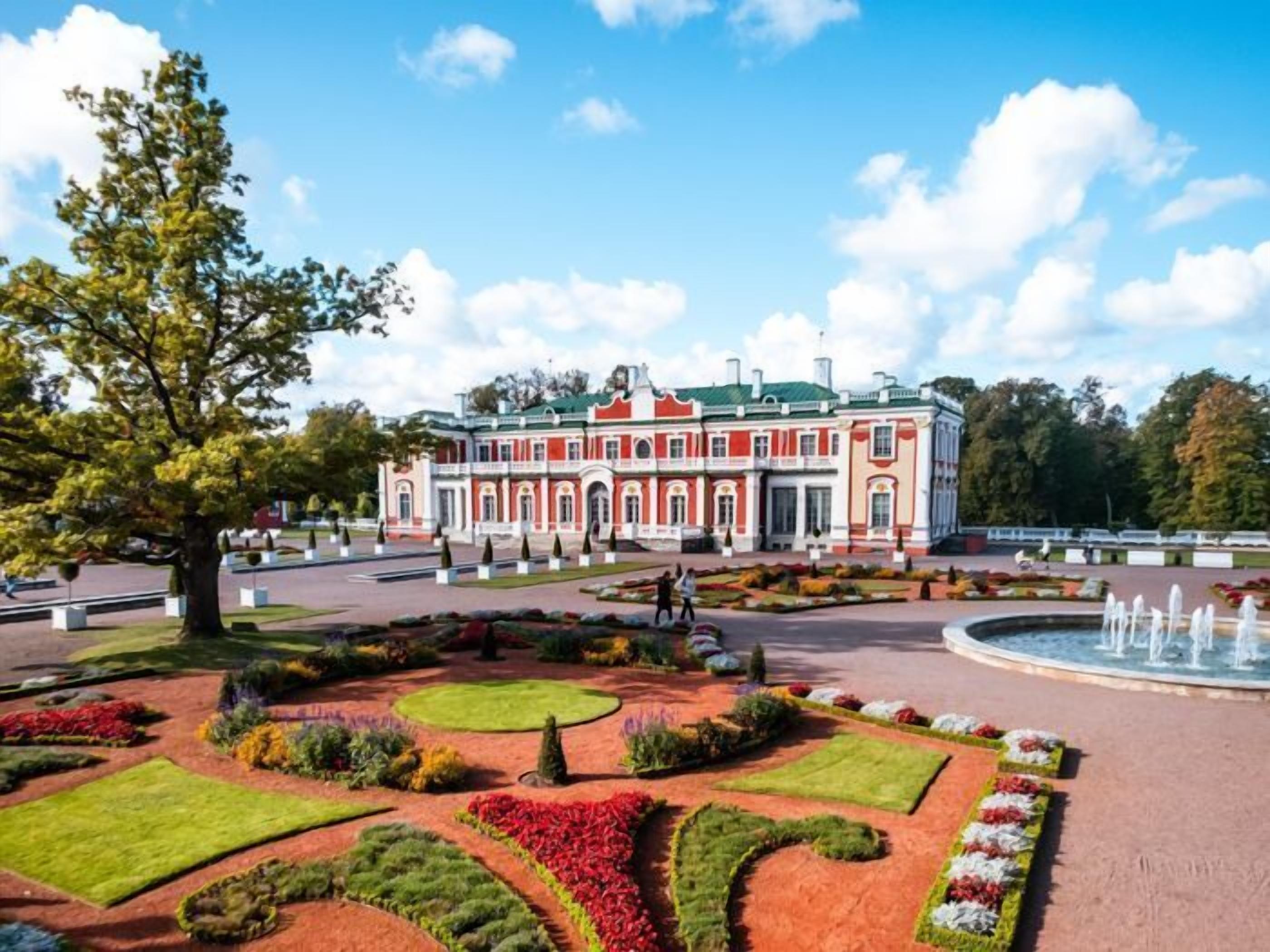 A large, historic building with a symmetrical facade, surrounded by a well-maintained garden with various flower beds and a central fountain.