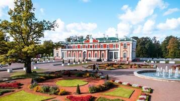 A large, historic building with a symmetrical facade, surrounded by a well-maintained garden with various flower beds and a central fountain.