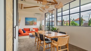 A modern dining area with a wooden table and chairs, a sofa with orange cushions, and large windows offering a view of buildings and greenery. - MariNara Lofts (Canggu)