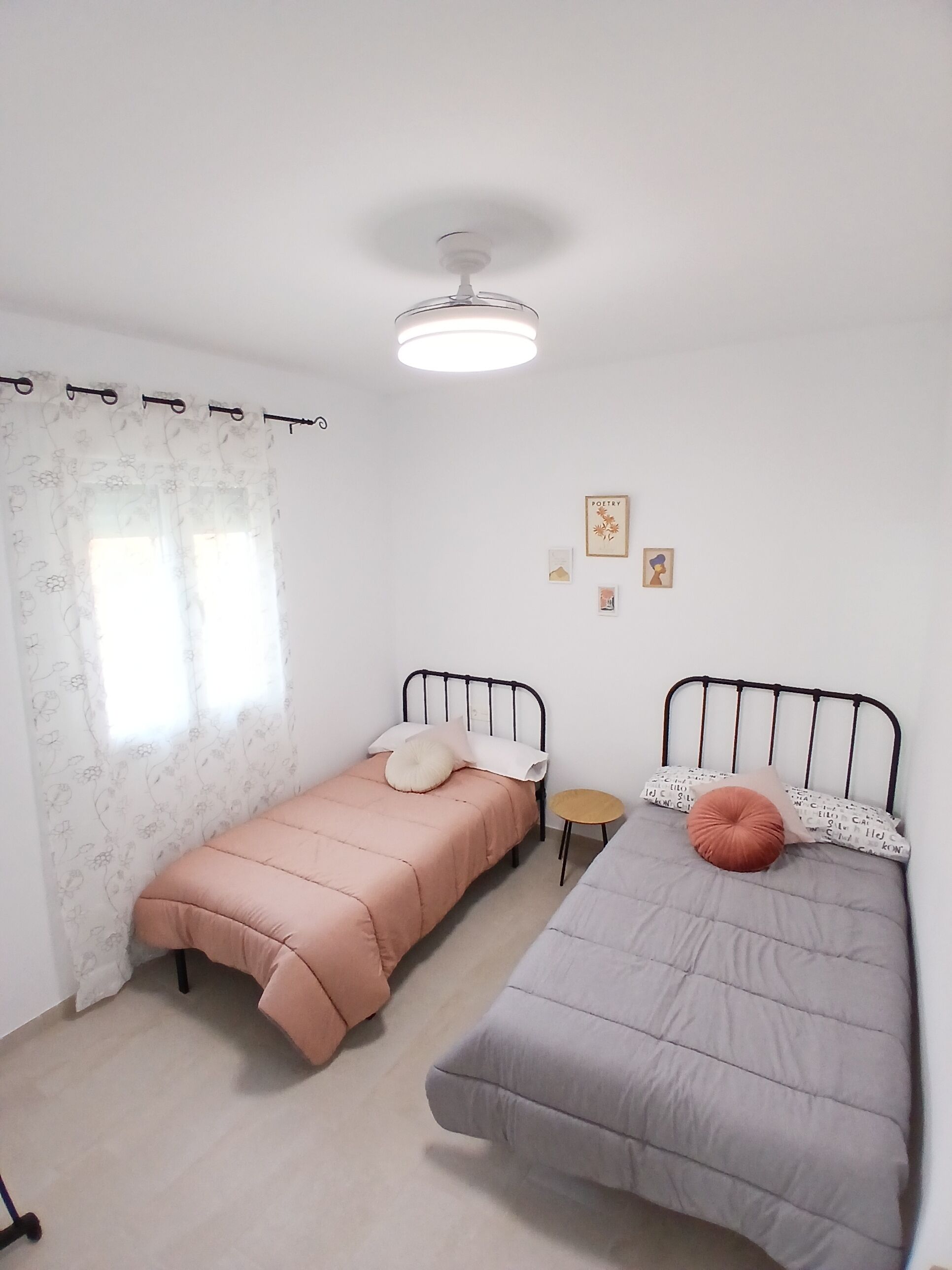 Una habitación pequeña y limpia con dos camas, una mesita y una lámpara en el techo.