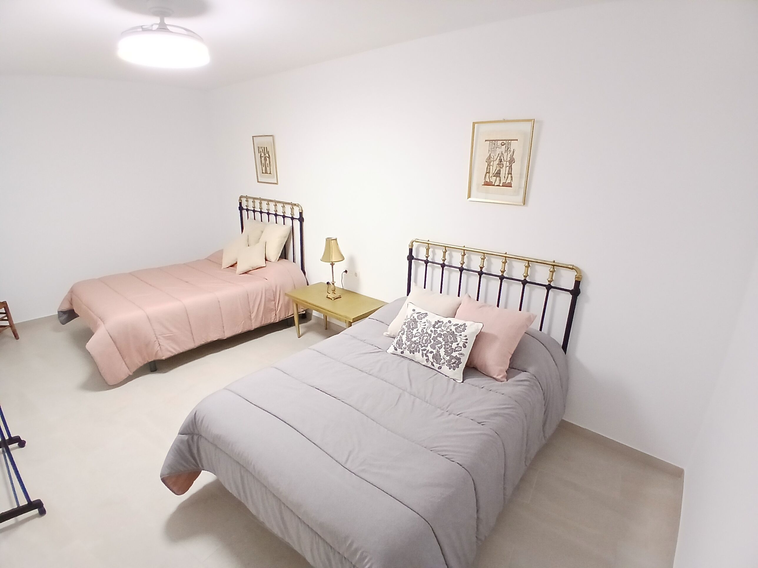 Quarto com duas camas, cada uma com cabeceira e almofadas, uma mesinha de cabeceira com abajur, e duas molduras com quadros na parede.