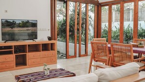 A living room with a TV, wooden furniture, and a view through large glass doors. - Ningaloo Villa (Gili Trawangan)