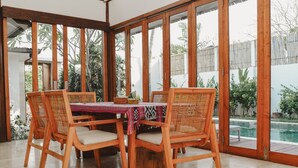 A dining area with a wooden table and chairs, large windows, and a view of a swimming pool. - Ningaloo Villa (Gili Trawangan)