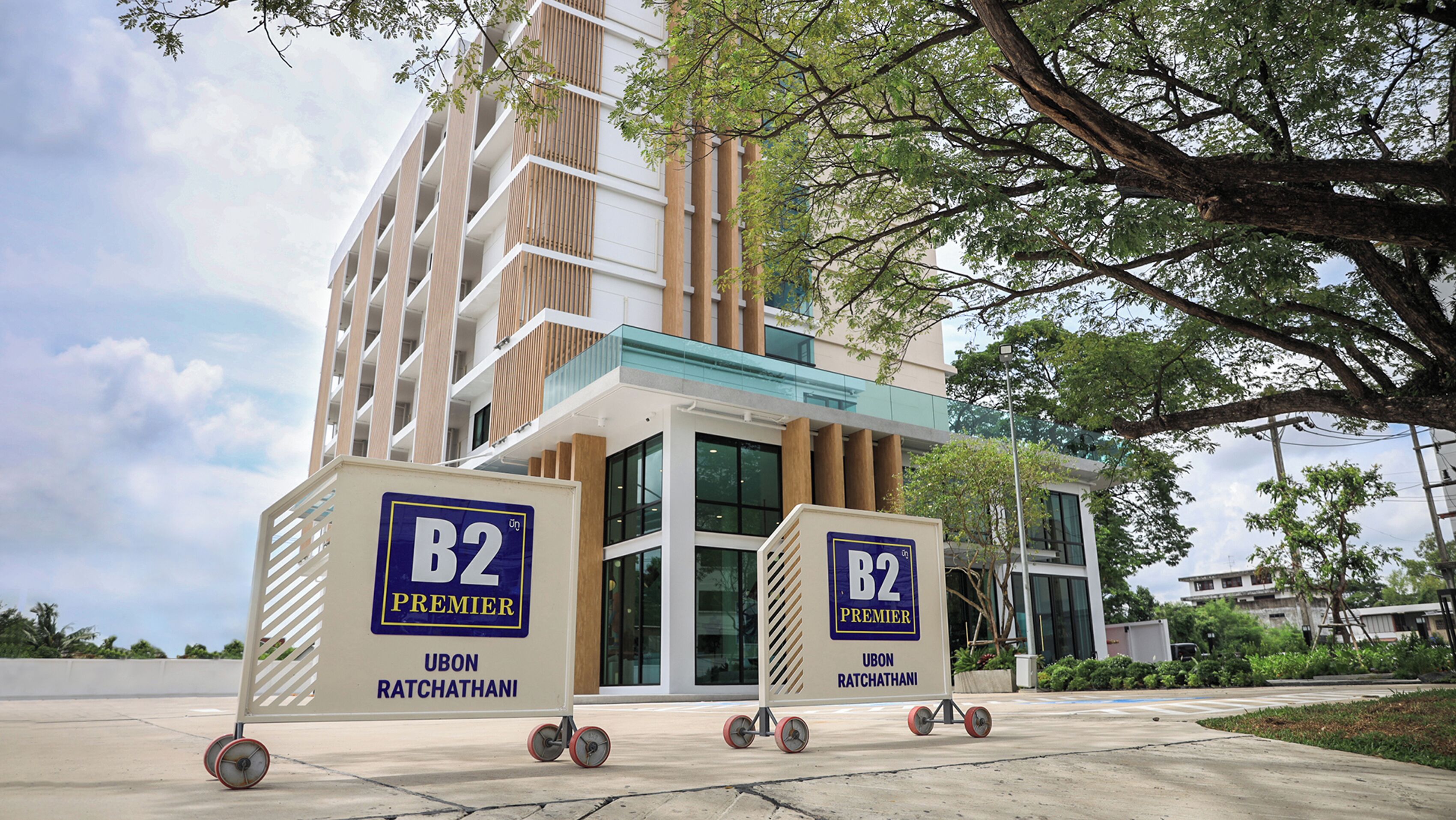 Two B2 Premier branded delivery carts outside a modern building with a glass entrance and a tree in the foreground.