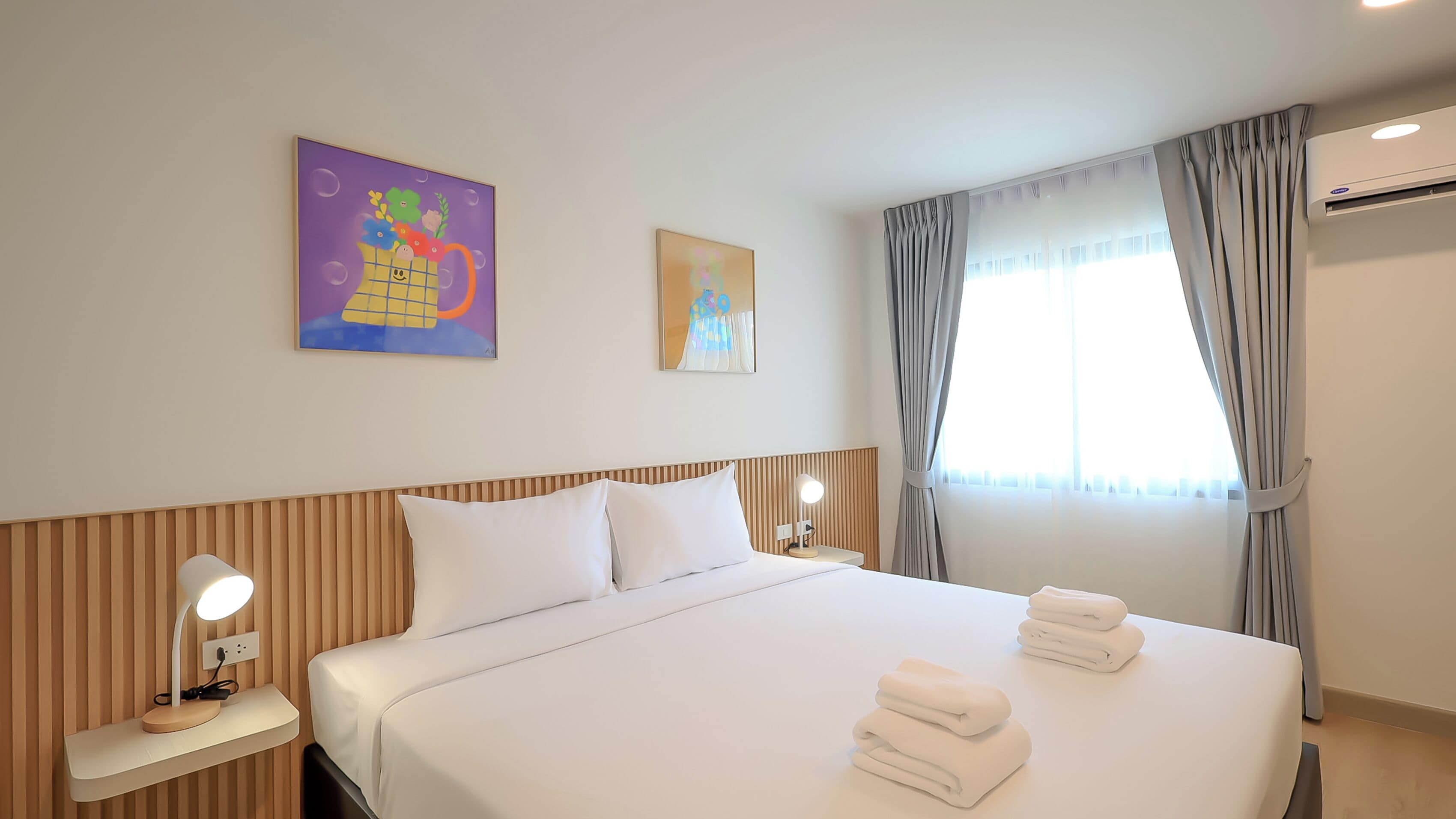 Una habitación de hotel con cama, dos mesitas de noche con lámparas, una ventana con cortinas y obras de arte en las paredes.