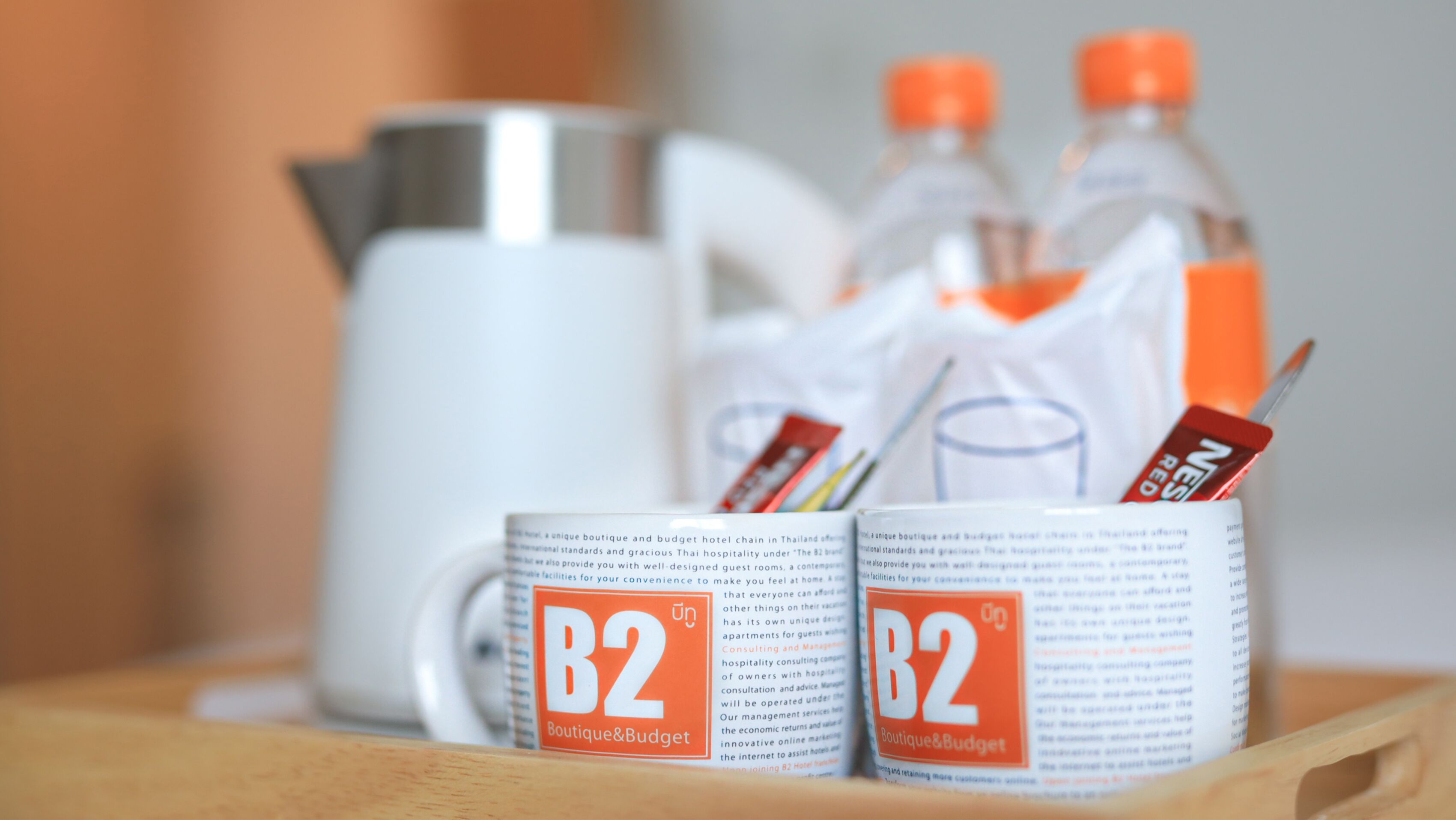 Two mugs with 'B2 Boutique & Budget' branding, a white coffee pot, and two bottles with orange caps.